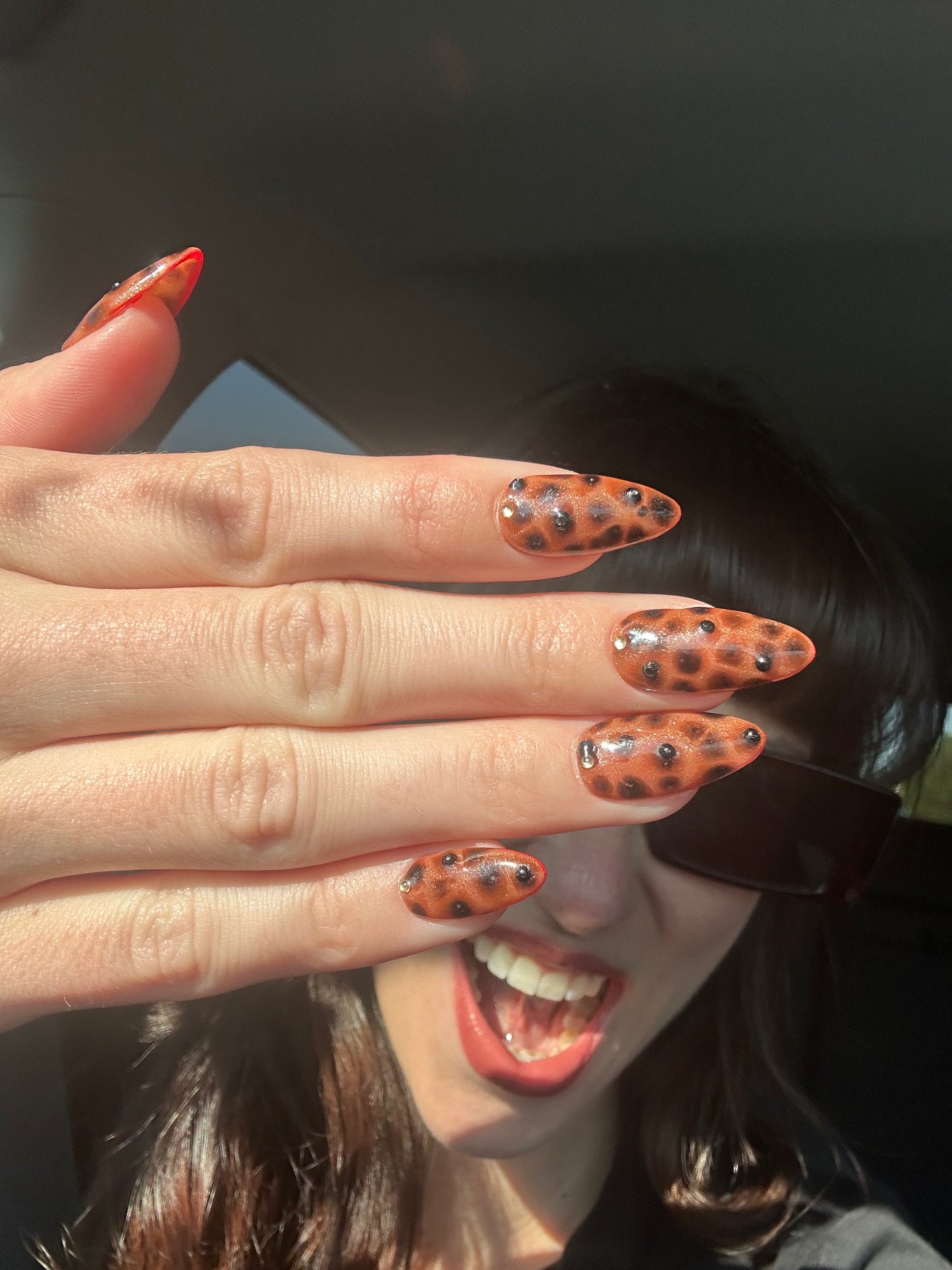 Close-up of a hand with orange nail polish and black polka dots, held over a blurred face.